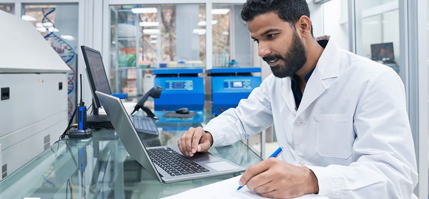 Biochemist in lab writing on laptop with notes on notepad