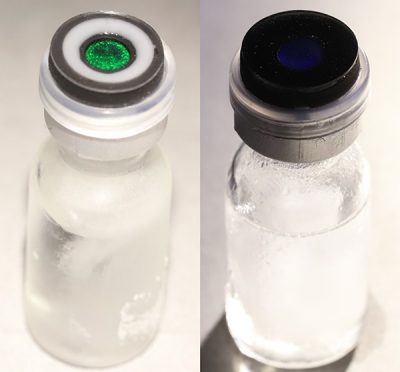 To the left, a vial filled with a colorless liquid has a label on the lid with a green-colored center dot surrounded by a white ring. To the right, a similar vial has a label that is completely black.
