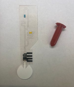 A photo of a small plastic device with a paper tab and printed circuit at one end next to an Eppendorf tube for scale. 